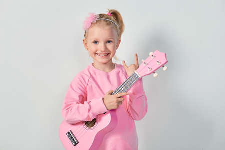 Cute caucasian little girl 4-5 year old in pink sweatshirt are playing the ukulele against gray...