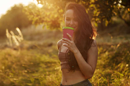 Young beautiful smiling woman in park and holding paper cup with coffee or tea on a tree and bush...