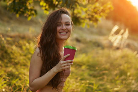 Young beautiful smiling woman in park and holding paper cup with coffee or tea on a tree and bush background in golden hour free space for textの写真素材