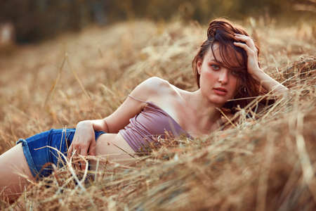 Beautiful sexy young woman posing in the hay of the sunset. Dressed in jeans shorts and beige top, lifestyle on nature. Romantic autumn mood, golden hourの写真素材
