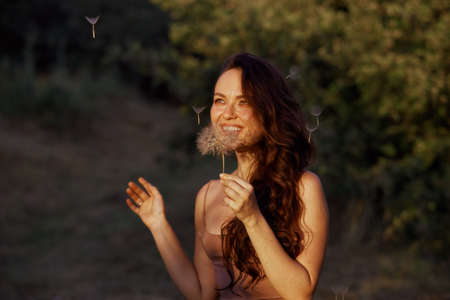 Beautiful happy woman blowing big dandelion. Outdoors, enjoy nature. Healthy smiling woman on summer lawn. Allergy free concept. Gorgeous slim caucasian, free space for textの写真素材