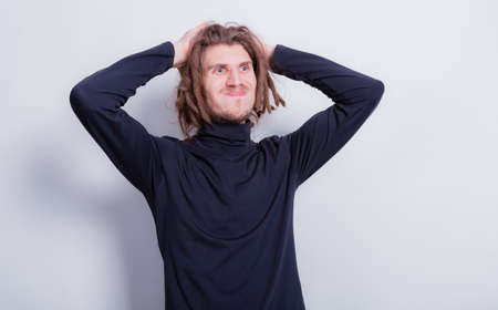 Surprised funny man holding his head, puffed out cheeks and compressed lips. Man in dark gray turtleneck and short dreadlocks on head hold hands to his head on a gray background copy spaceの写真素材