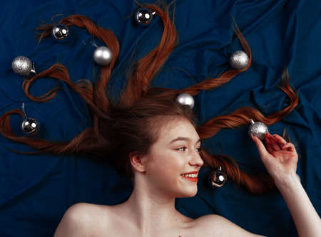 Cute redhead young woman lying on blue bed with of decorative christmas balls on ends of her hair. Top viewの写真素材