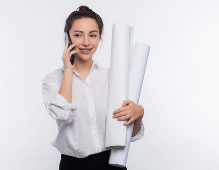 Young and cute business woman holds blueprints in her hand and use her phone. She is happy and shares the news on the phone her project was approved and she got the job isolated on white copy spaceの写真素材