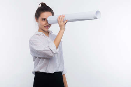 In search of new projects concept. Young and cute business woman holds blueprints in her hands like a telescope isolated on white copy spaceの写真素材
