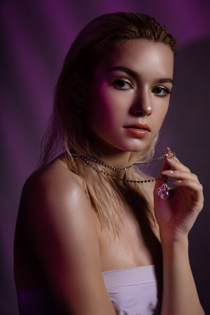 High fashion model woman face in colorful bright neon purple lights. Beautiful woman posing in the studio and holding or showing a chain, glowing glitter bright neon makeup.の写真素材