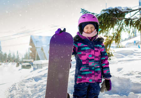 Little cute girl snowboarding at ski resort in sunny winter day. Caucasus Mountains. Portrait of kid in helmet and snowboard in winter resortの写真素材