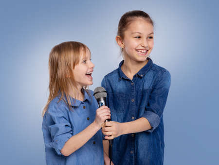 Two little funny children girls 6 and 8 years old sing a song in a microphone. The concept is childhood, lifestyle, music, singing, friendship on blue backgroundの写真素材