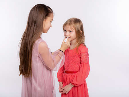 Two girls friends or sister applying lipstick on lips. Helping each other doing make-up to prepare for the holiday. Together having fun isolated over white backgroundの写真素材
