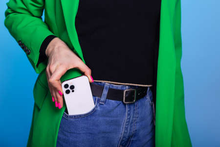 Young woman takes out or puts in a pocket mobile phone. Woman wears green blazer and blue jeans. Smartphone with three cameras in front pocket on blue background. Mockup for print or design templateの写真素材