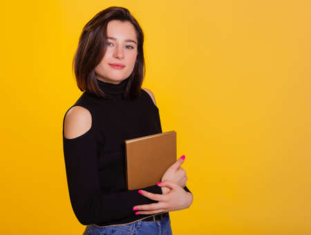 Pretty woman with a book in hands smiling at camera. Clever and excited lady embrace book. Studio shot isolated on yellow backgroundの写真素材
