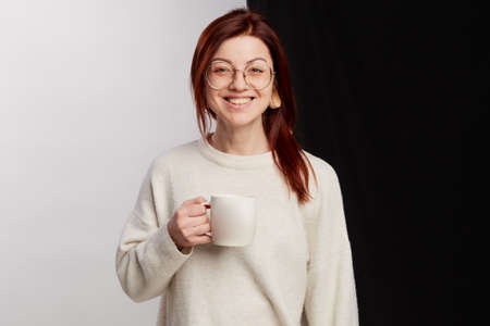 A hot drink invigorates. Redhead woman holding cup of tea or coffee in hand, face expression stands...
