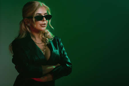 Studio fashion shot of the beautiful rock woman dressed in black leather jacket and black body lingerie, sunglasses on the face. Seductive lady in green light on a dark green backgroundの写真素材