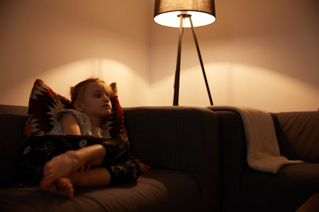 Beautiful little girl sitting on cosiness sofa in the living room at home and watching TVの写真素材