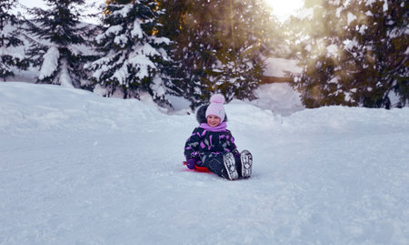 Little girl enjoying the winter sledding time. Child playing and having fun riding on a snowy hill. Slide down from snow slope sitting in plateの写真素材