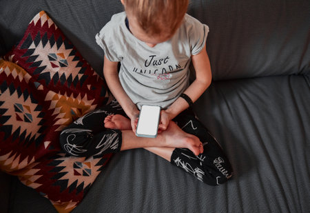 Cute child girl sitting on sofa in living room use smartphone and play online games. Holding phone and looking on screen device. Kid sitting on the couch and rest in living room. Technology concept.の写真素材