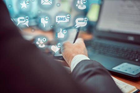 A young businessman works in a new modern office at a table on a laptop and holds a mobile phone in his hands. IT sphere of life concept.の写真素材