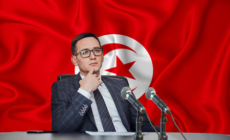 Young man in glasses and a jacket at an international meeting or press conference negotiations, on the background of the flag Tunisiaの写真素材