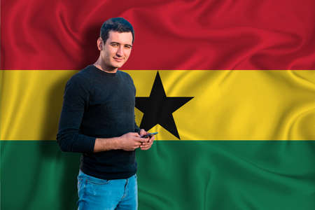Ghana flag on the background of the texture. The young man smiles and holds a smartphone in his hand. The concept of design solutions.の写真素材