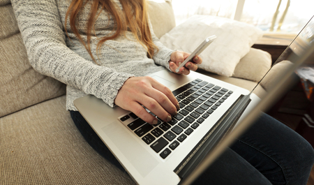 The young woman is sitting on the sofa and working at laptop and holds phoneの写真素材