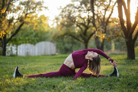 Split-leg on sunset. Girl in red sportswear doing yoga pose in the autumn park. Balance, fitness, stretching and relaxationの写真素材