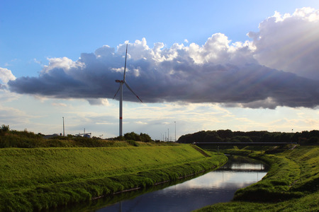 A wind turbine in a peaceful landscape with green grass, blue skye and a river