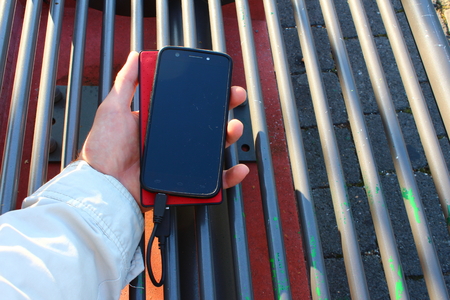 A black smartphone and a red powerbank in a male hand on the benchの写真素材