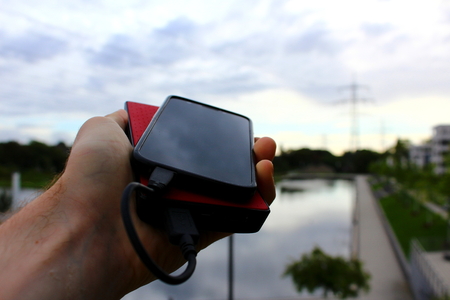 A black smartphone and a red powerbank in a male hand in a park with a pondの写真素材