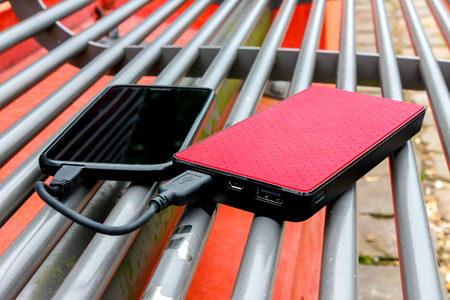 Black smart phone and red power bank battery charging on park benchの写真素材