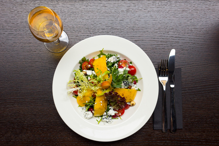 Dessert salad with mango and goat cheese, glass of orange wine and fork and knife on black wooden table background, top viewの写真素材