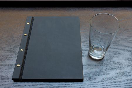 Black or dark grey restaurant menu with copy space and empty glass on dark wooden table backgroundの写真素材
