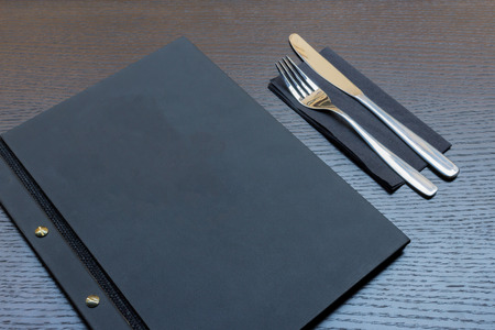 Black or dark grey restaurant menu with copy space and fork and knife on dark wooden table backgroundの写真素材
