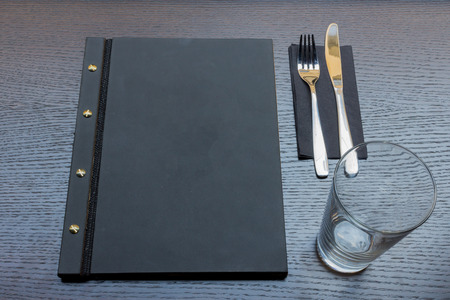 Black or dark grey restaurant menu with copy space and fork and knife and empty glass on dark wooden table backgroundの写真素材