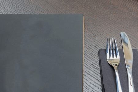 Black or dark grey restaurant menu with copy space and fork and knife on dark wooden table background closeupの写真素材