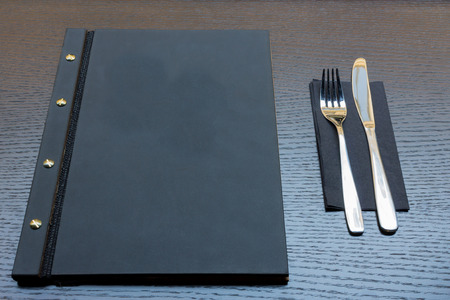 Black or dark grey restaurant menu with copy space and fork and knife on dark wooden table backgroundの写真素材