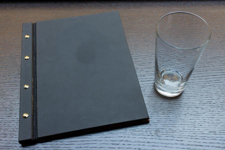 Black or dark grey restaurant menu with copy space and empty glass on dark wooden table backgroundの写真素材