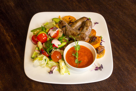 Delicious fried sausages and vegetable salad in a white dish with red sauce on brown table backgroundの写真素材