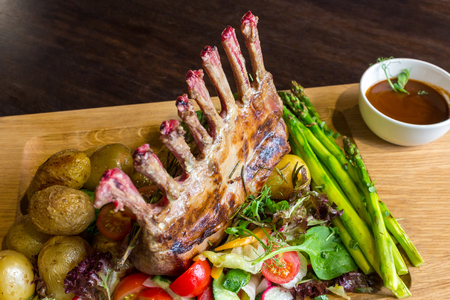 Delicious lamb meat crown roast with vegetable salad and brown sauce on wooden platter on brown wooden tableの写真素材