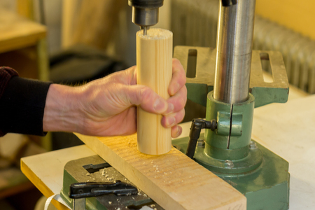 Worker hand holding a wooden detail and processing it with a drilling machine on a work bench in a workshopの写真素材
