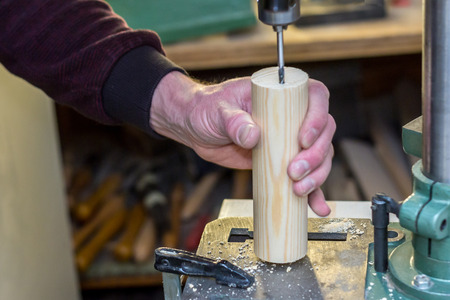 Labourer hand holding a wooden detail and processing it with a boring machine on a work bench in a workshop with instruments in the backgroundの写真素材
