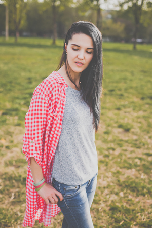 Beautiful female hipster in red plaid shirt, grey t-shirt and ripped blue jeans in a casual style posing in green parkの写真素材