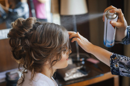Pretty young bride preparing for wedding at home in the morning. Stylist making hairstyle and apply hairspray on curly hairの写真素材