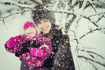 happy young mom in fur coat with little daughter in pink pattern coat smiling, hugging and having fun in snowfall in the winter park. Concept of happy familyの写真素材