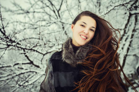 portrait of beautiful young woman with long brunette hair in fur coat smiling and spending time outdoors in the winter forest.の写真素材