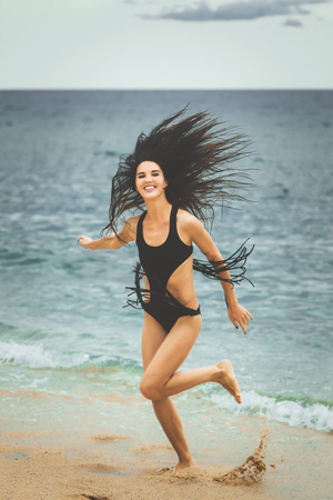 Young beautiful playful teen on the beach, ocean and mountain background. She is dressed in black swimwear. hipster style, soft lightの写真素材