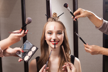 Beautiful young model with professional unusual makeup and a coiffure with topknot and braid made by stylists in a beauty salon is surrounded by hands holding brushes and palette.の写真素材
