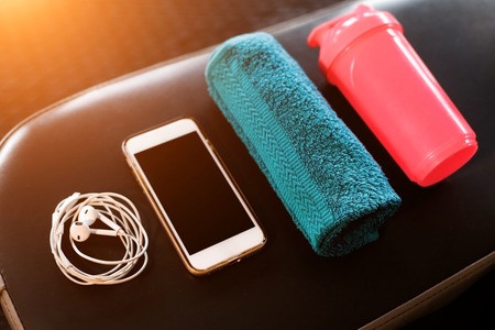 set of mobile phone, earphones, blue towel and a pink shaker lying on a black leather bench in a fitness center. sport equipment and gadgets conceptの写真素材