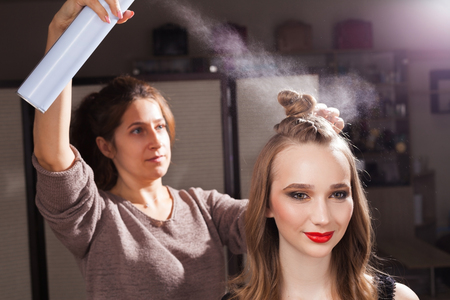 hairdresser fixing a coiffure with an unusual bun of a young beautiful girl using a hair spray in a beauty salon. concept of professional stylist studyingの写真素材