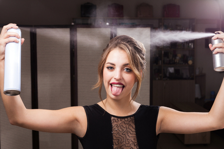 beautiful happy joyful model with perfect makeup and coiffure is fixationing her hair by two bottles of sprays. girl is showing a tongue. concept of professional hairdresser productsの写真素材