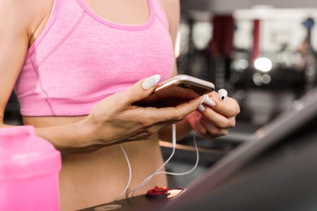 slim girl holding a smart phone and earphones choosing music for exercising. woman standing by the...
